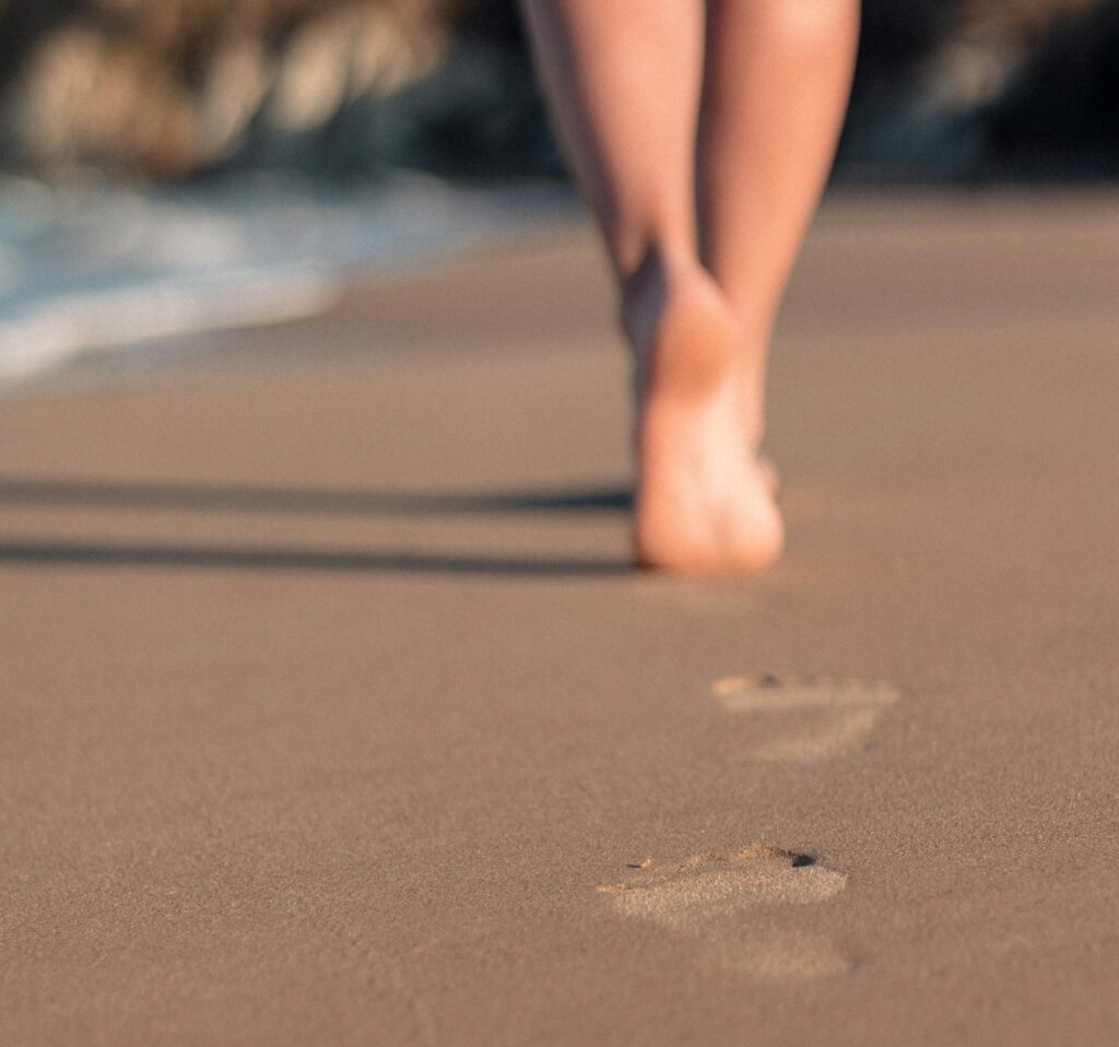Close-up of a woman's legs walking barefoot on a sandy beach, leaving footprints behind.